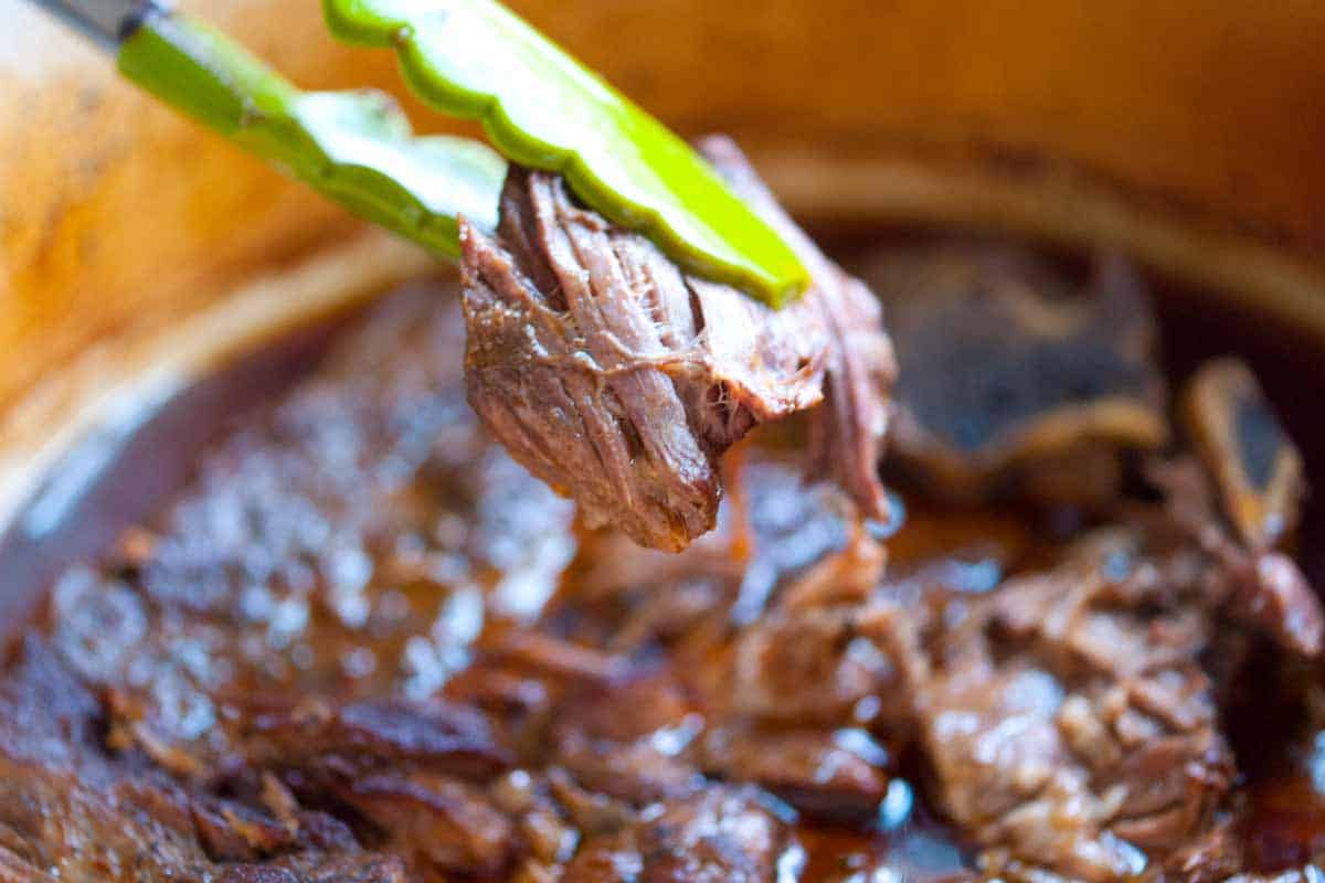 How to Make Irresistible Shredded Beef Tacos Tongs pulling up super tender shredded beef from a pot