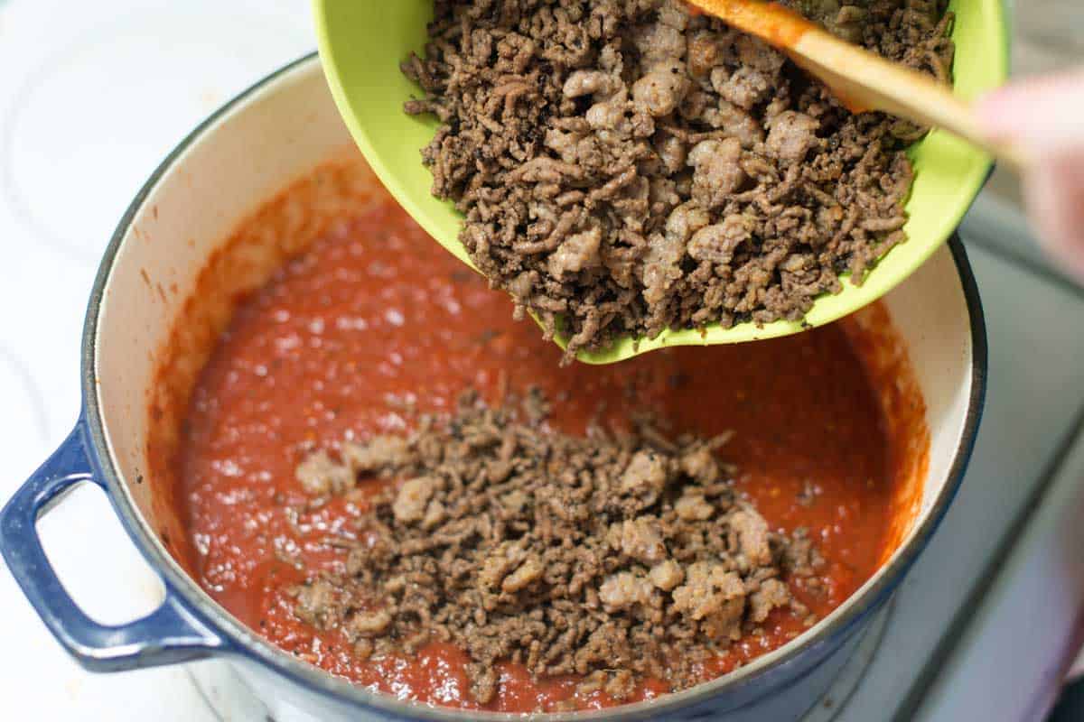 Stirring browned sausage and ground beef into the lasagna sauce