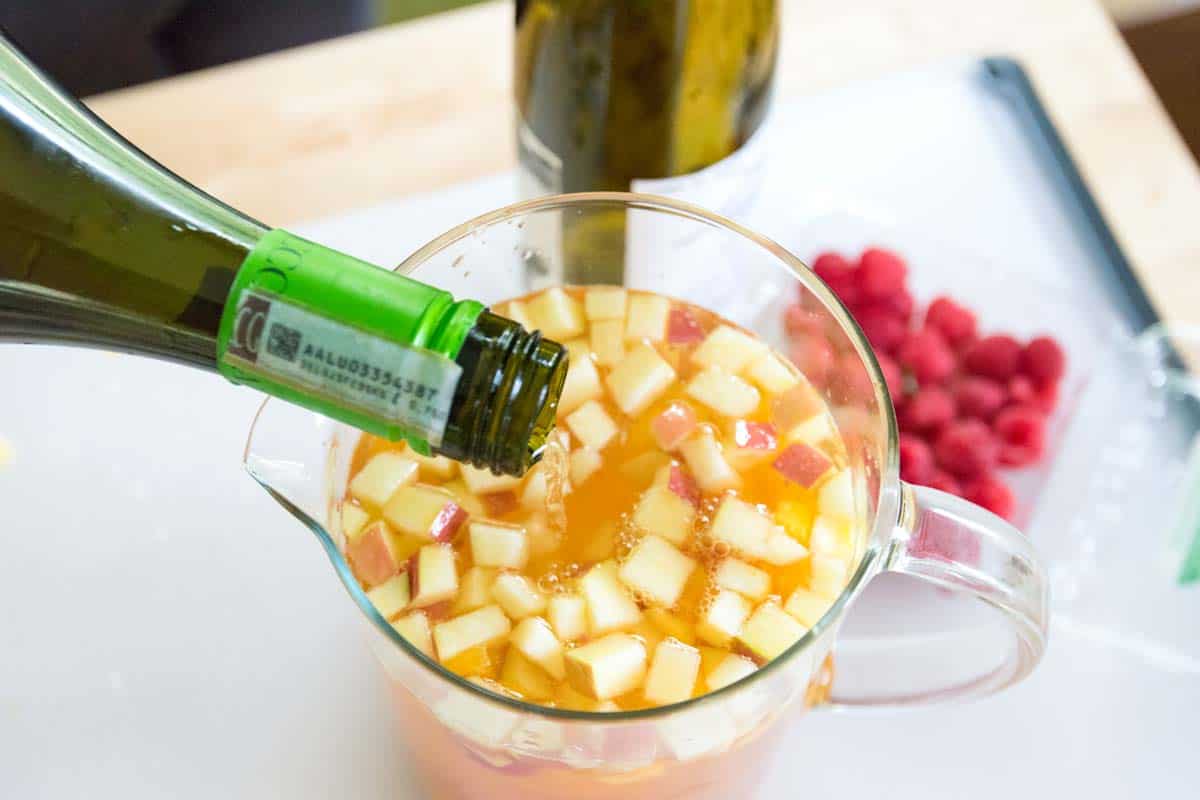 Pouring white wine into a pitcher with apples, peaches, and raspberries