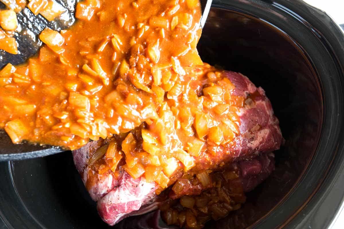 Adding onions, garlic, tomato paste, and spices on top of pork in a slow cooker