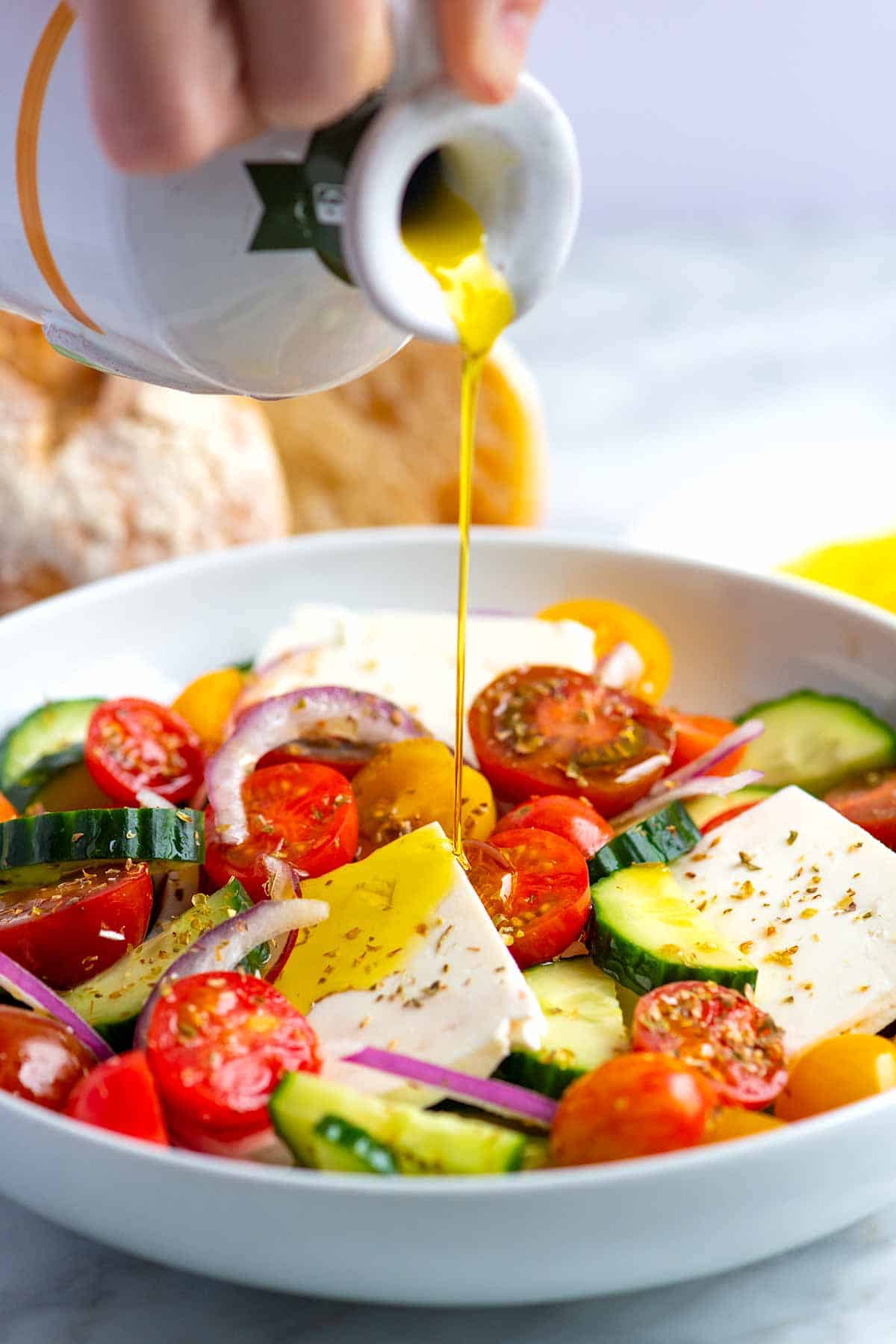 Olive oil being poured over tomatoes, cucumber, feta, and onion for Greek salad