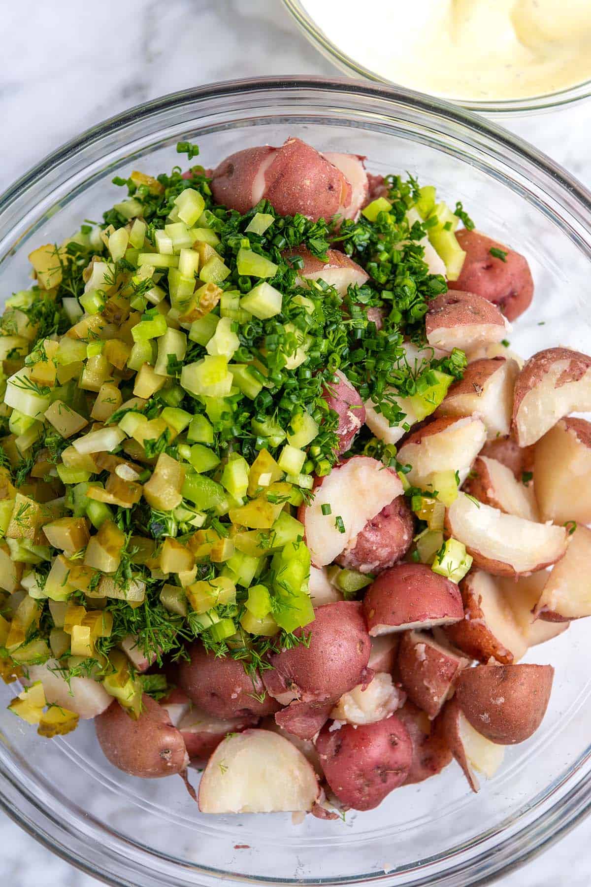 Mixing bowl with red potatoes, herbs, chopped celery, and chopped pickles.