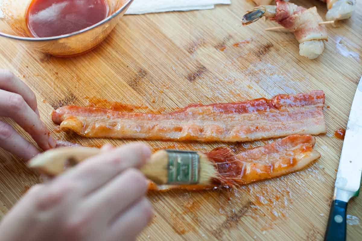 Brushing par-baked bacon with spicy maple glaze before wrapping the shrimp