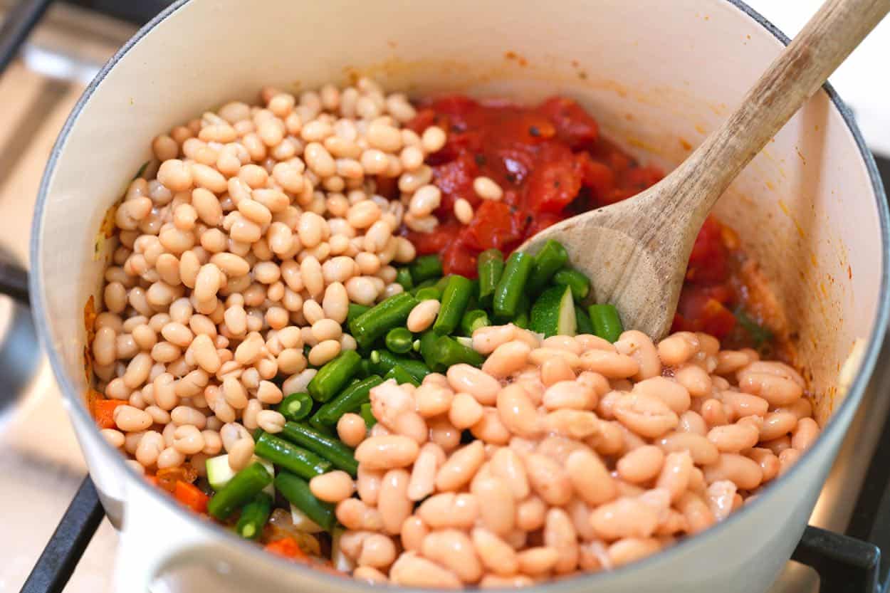 Adding beans, green beans, tomatoes, and zucchini to a minestrone soup pot.
