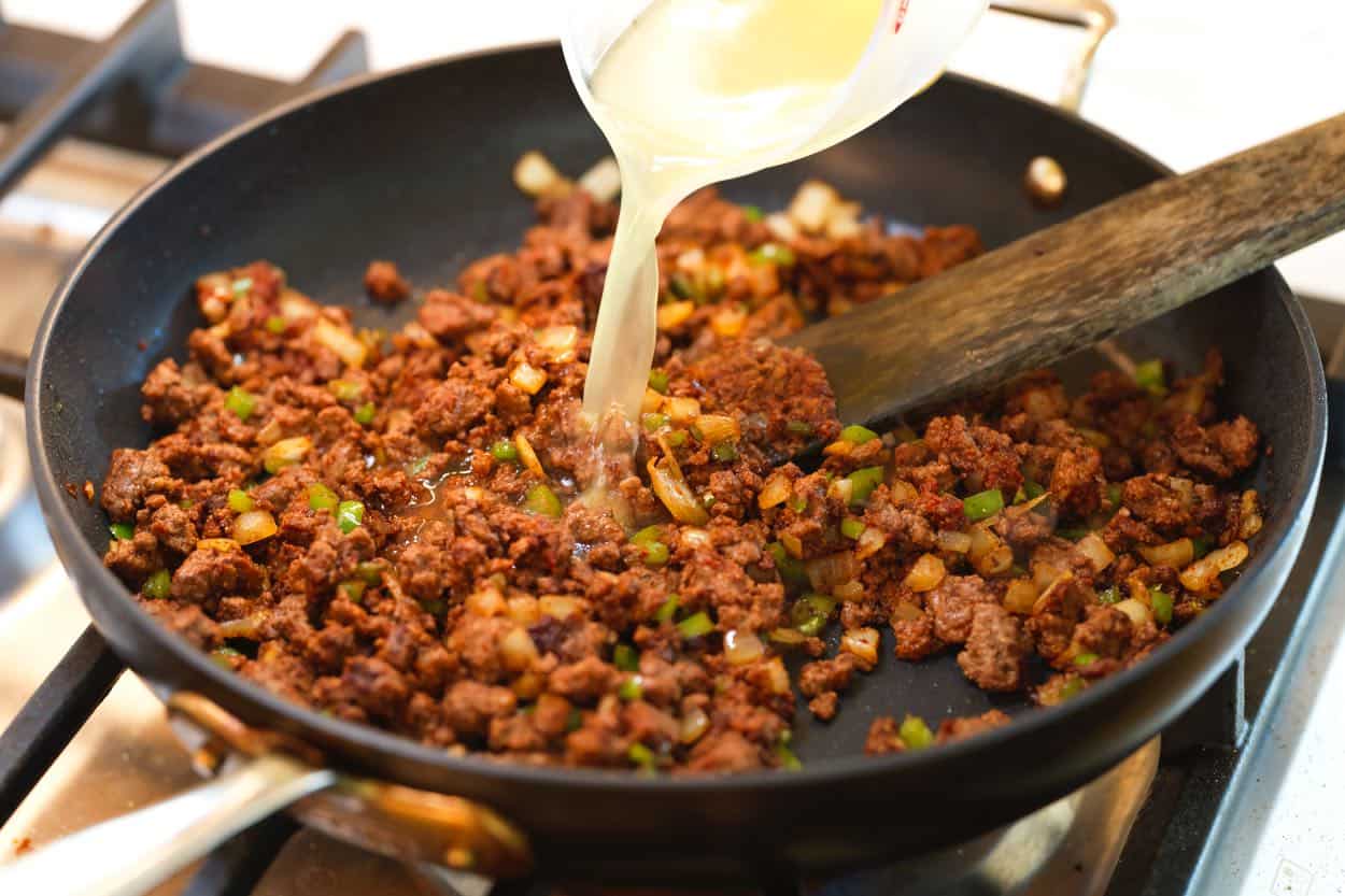 Pouring stock into a skillet with spiced ground beef for taco meat