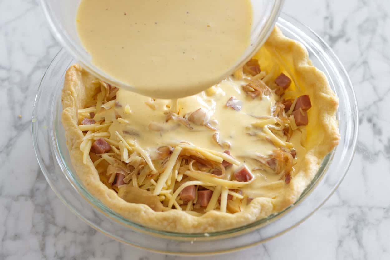 Pouring custard into a pie crust filled with ham and cheese for quiche