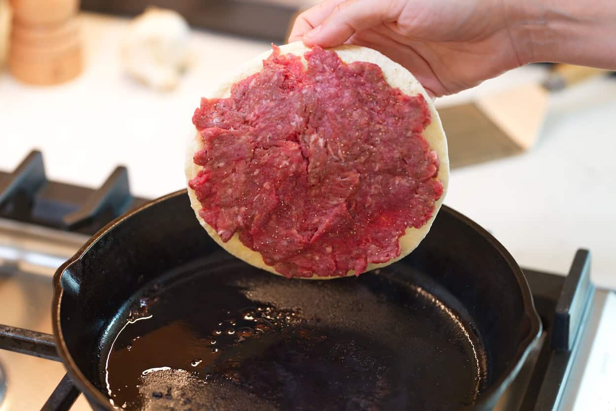 Placing a tortilla with ground beef spread over it into a pan for smash tacos