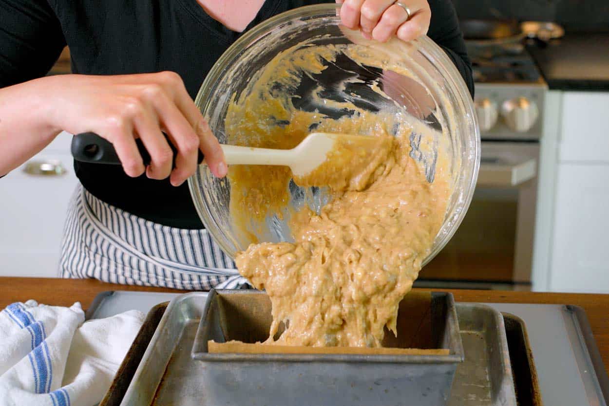 Spooning banana bread batter into a loaf pan sitting on two baking sheets stacked on top of each other.